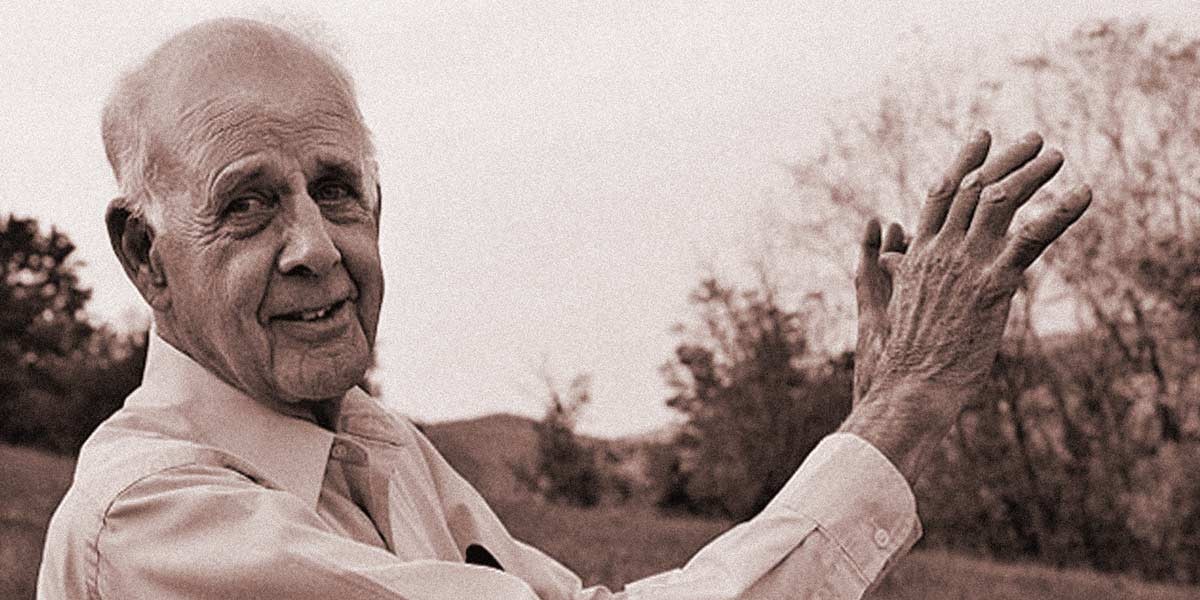 Elderly man in a light shirt outdoors, smiling and clapping with trees and hills in the background.