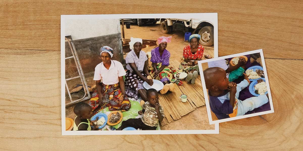 Four women and two children in colorful clothing sitting outdoors eating food from plates on a mat.