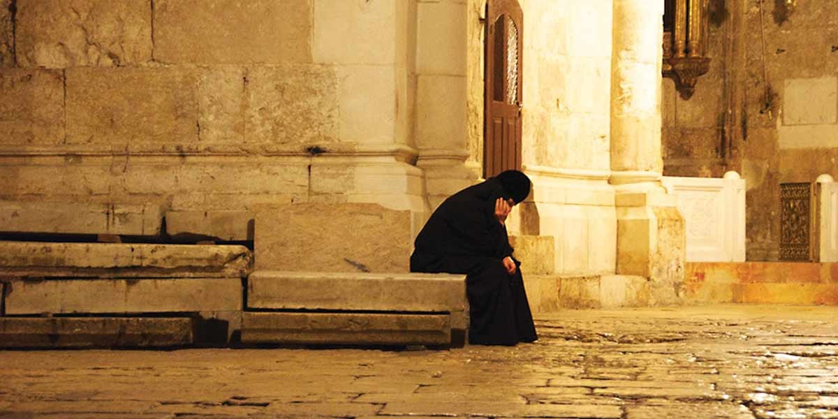 Person draped in black sitting alone on stone steps beside a historic stone building at night.