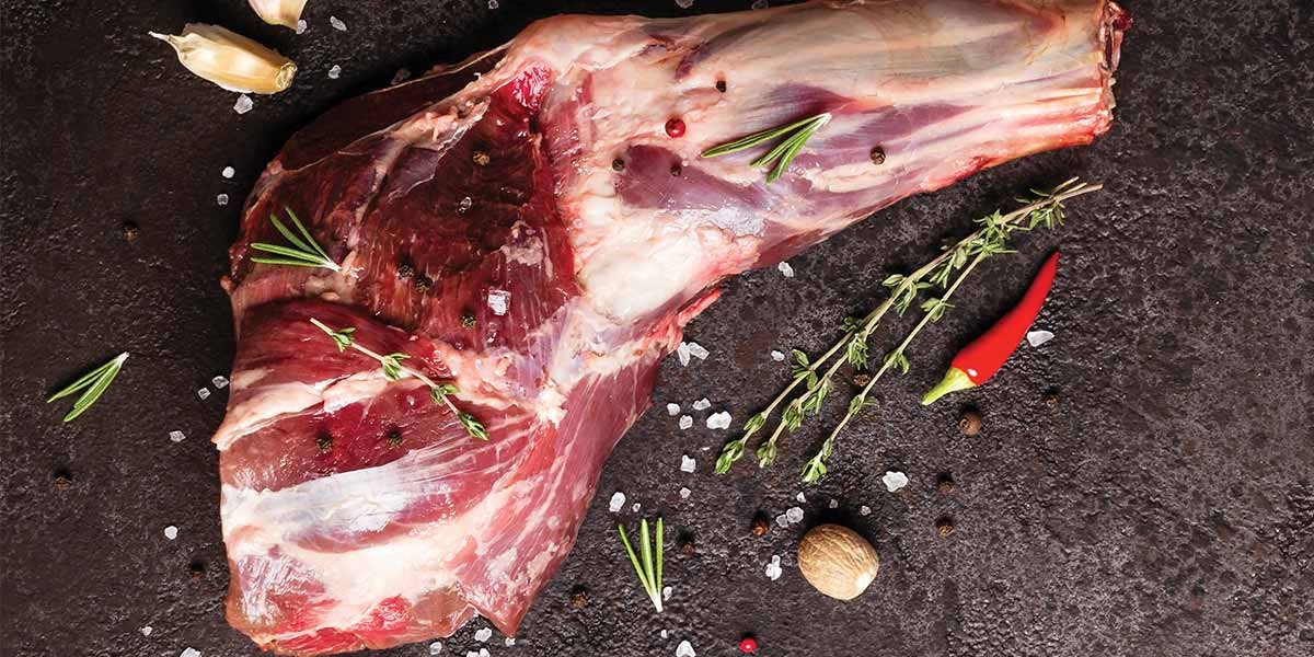 Raw leg of lamb on a dark surface with garlic, herbs, chili, peppercorns, and coarse salt.