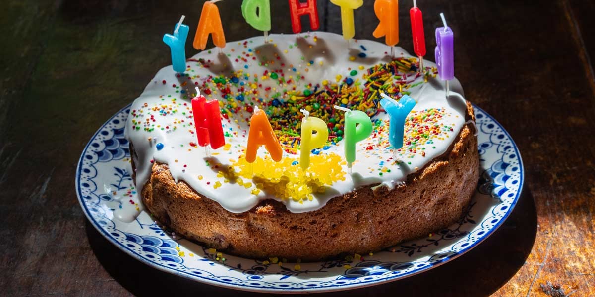 Round birthday cake with white icing, colorful sprinkles, and letter candles spelling HAPPY BIRTHDAY.