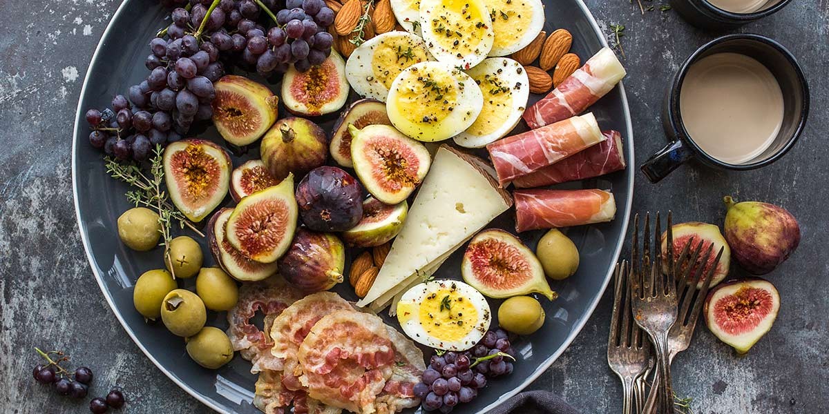 Plate with figs, grapes, boiled eggs, cheese, almonds, olives, prosciutto, and bacon on a dark tabletop.