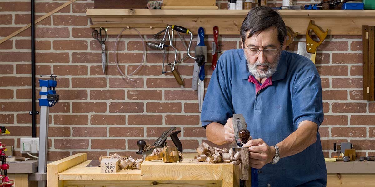 Man in blue shirt using hand planer on wood at a workbench in a woodworking shop.