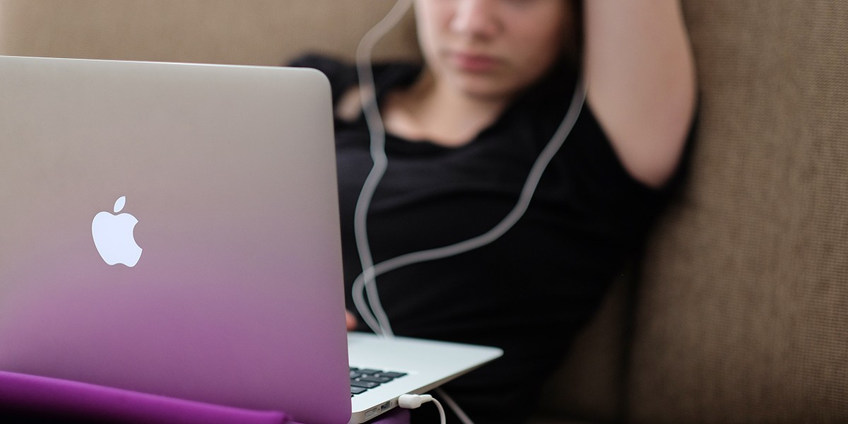 Person with earphones using a silver Apple laptop while sitting on a couch.