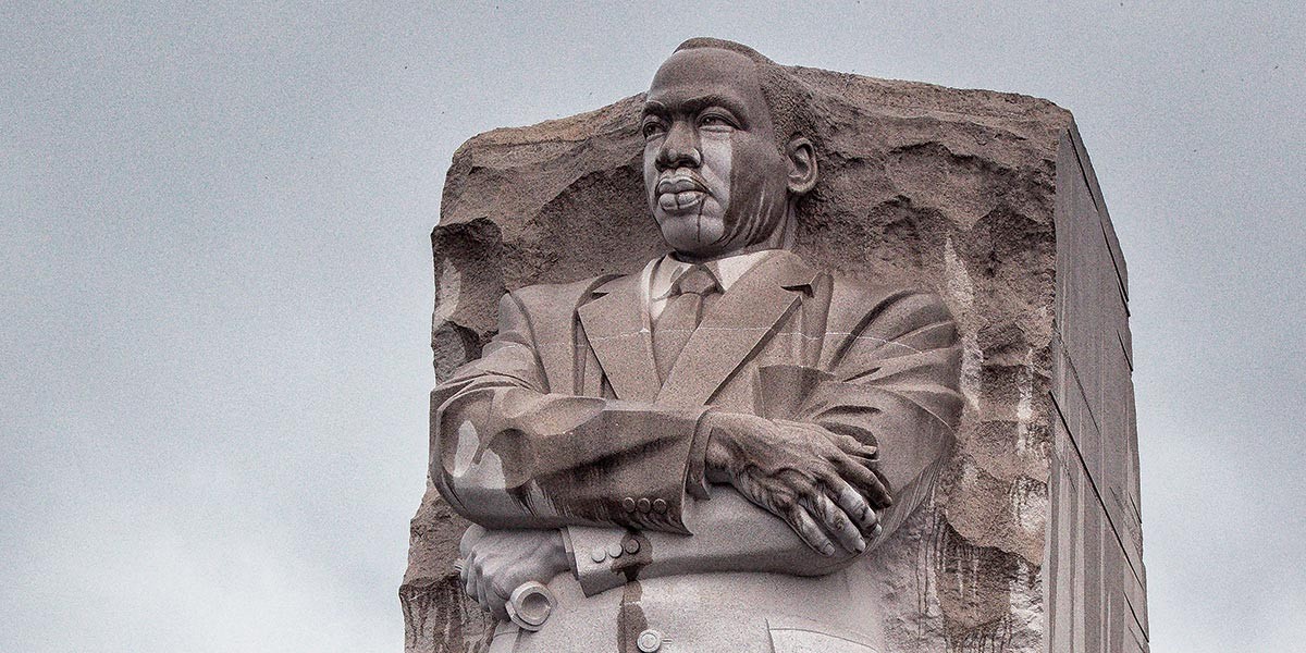 Statue of Martin Luther King Jr. carved in stone with arms crossed against a cloudy sky.