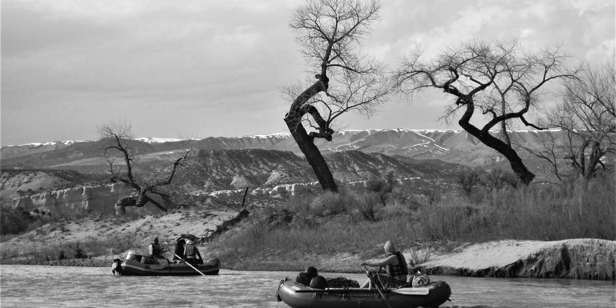 Two people rafting on a river with leafless trees and mountains in the background.