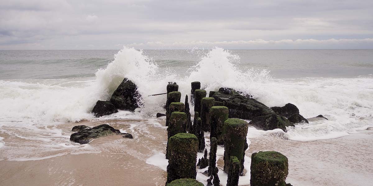 Ocean waves crashing against moss-covered wooden posts and rocks on a sandy beach under cloudy sky.
