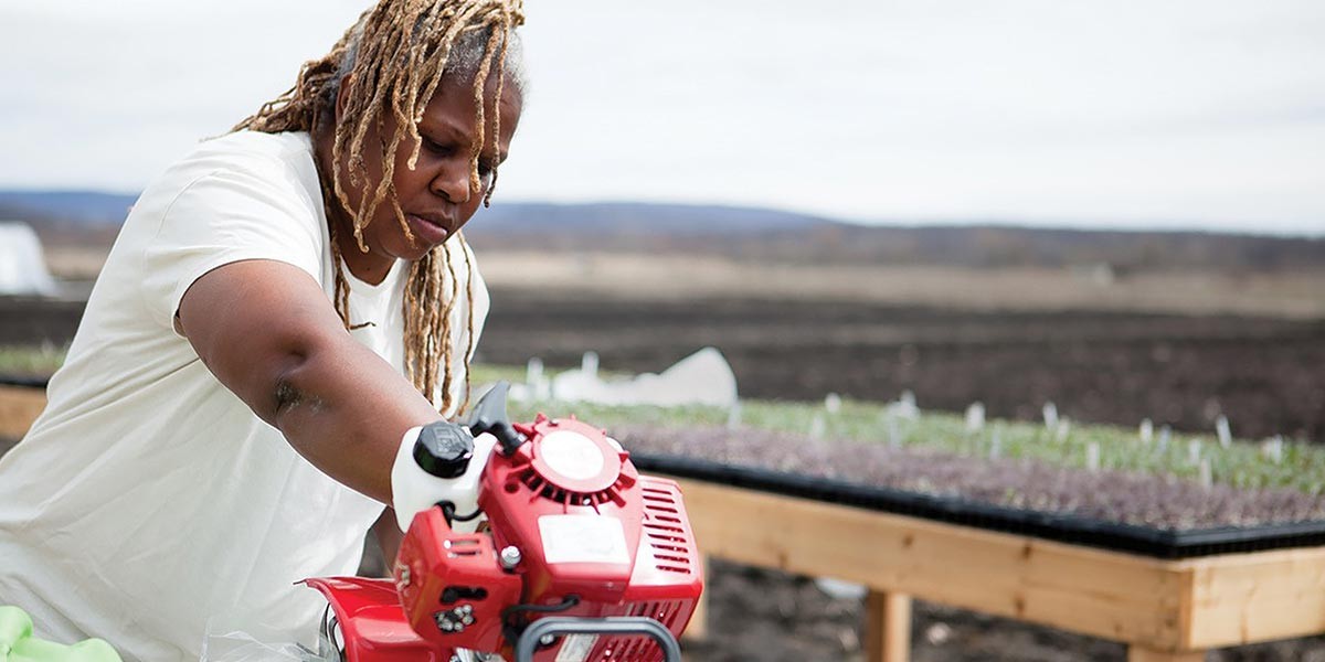 Person with blond dreadlocks using a red motorized gardening tool near seedling trays outdoors.