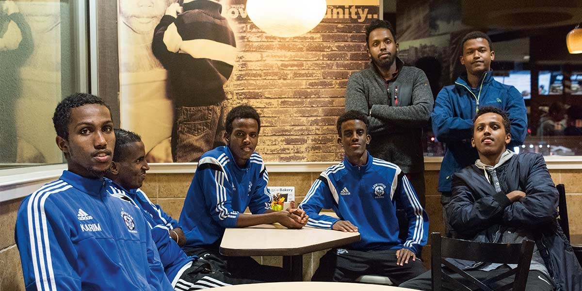 Group of young men, some in blue sports jackets, sitting and standing inside a casual dining space.