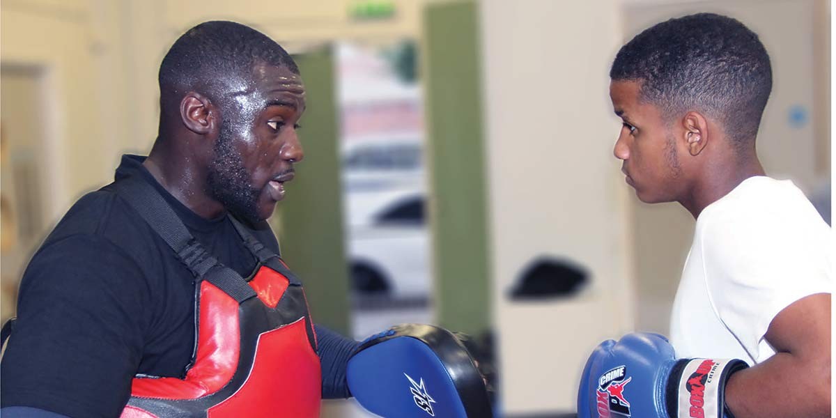 Two men facing each other wearing boxing gloves and headgear in a gym setting.