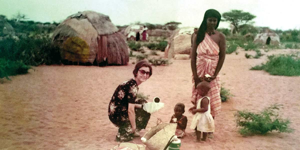 A woman in sunglasses crouches near two children and a standing woman in a sandy rural setting with huts.