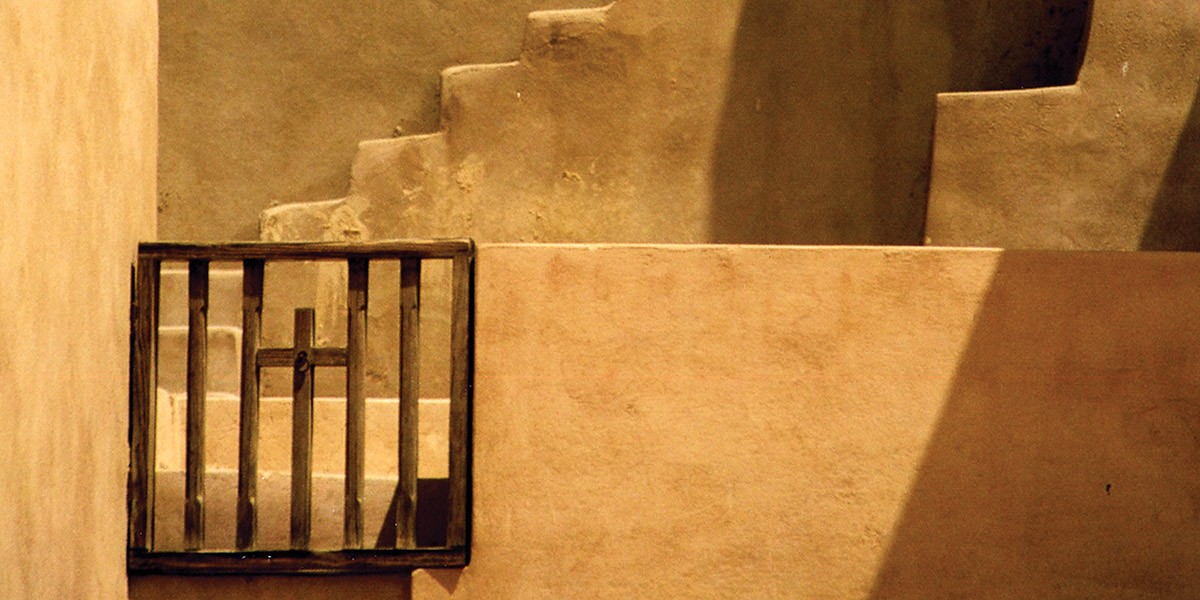 Wooden gate with a cross in front of beige adobe-style stepped walls bathed in sunlight and shadow.
