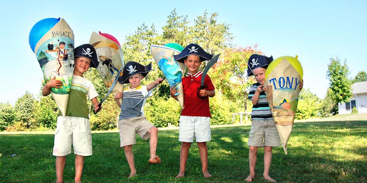 Four young boys wearing pirate hats, holding decorated cones and plastic swords outdoors on grass.