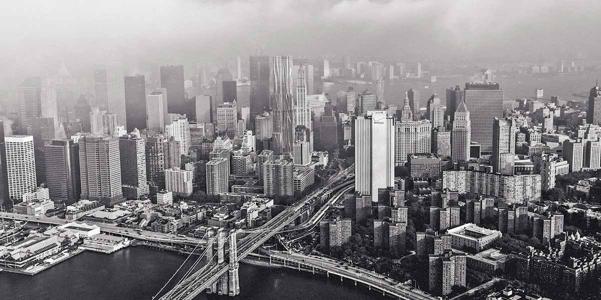 Black and white aerial view of a river, bridge, and dense cityscape with skyscrapers in foggy weather.