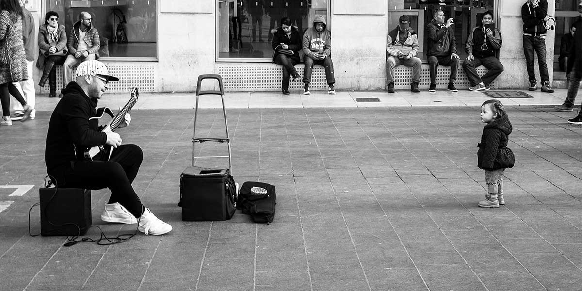 Black and white photo of a street musician playing guitar while a toddler stands and watches nearby.