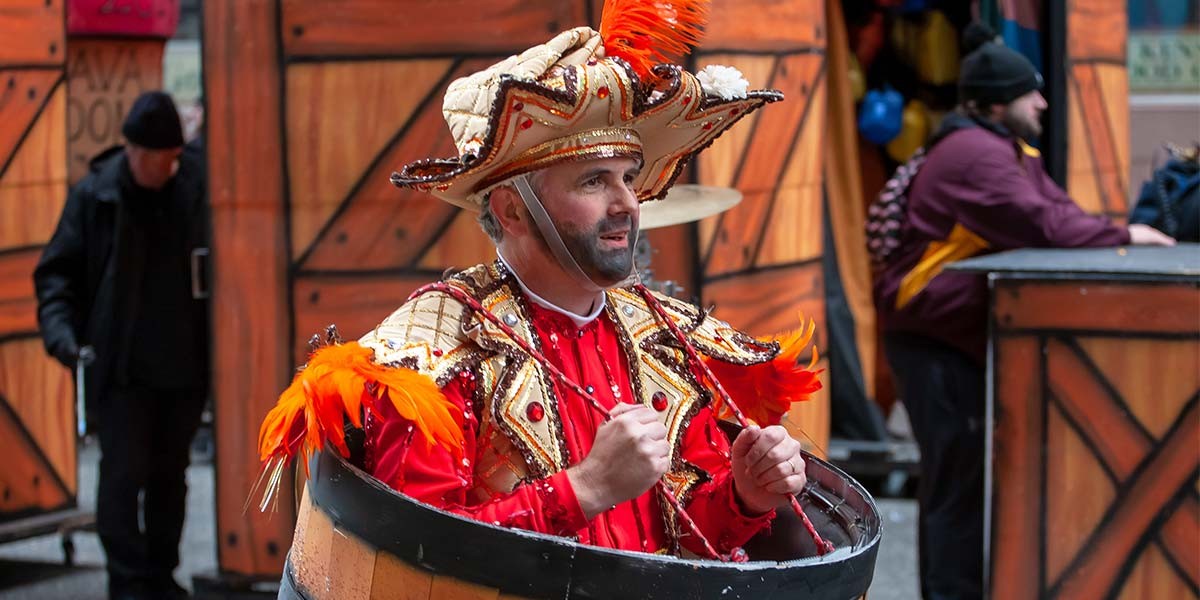 Man dressed in a colorful costume with a large hat and orange feathers sitting in a wooden barrel.