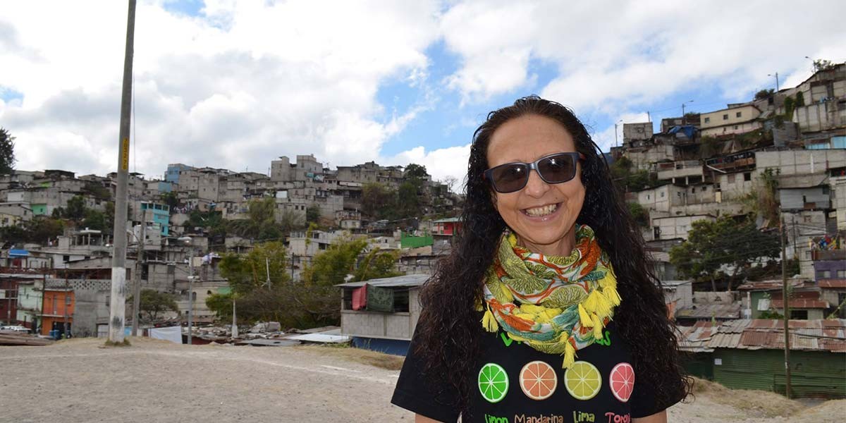 Smiling woman with sunglasses and colorful scarf in front of a hillside neighborhood with dense housing.