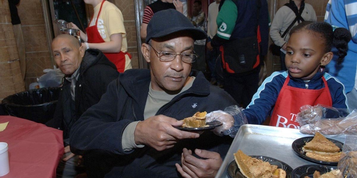 Young girl wearing red apron handing a plate with pie to a seated man in dark hat and glasses.