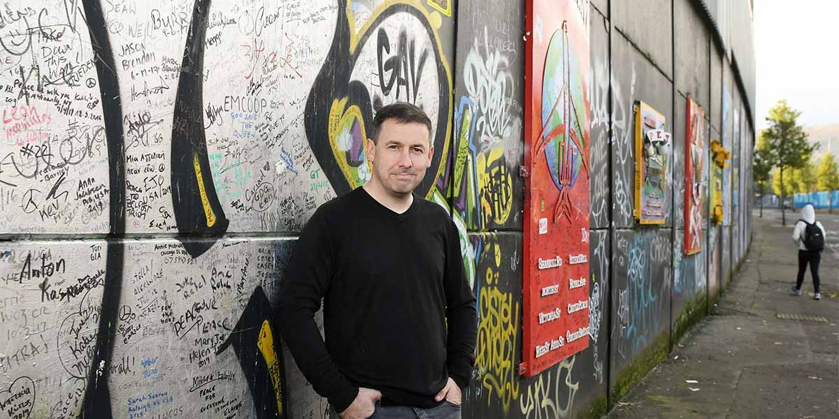 Man in black shirt standing by a graffiti-covered wall with signatures and colorful art panels.