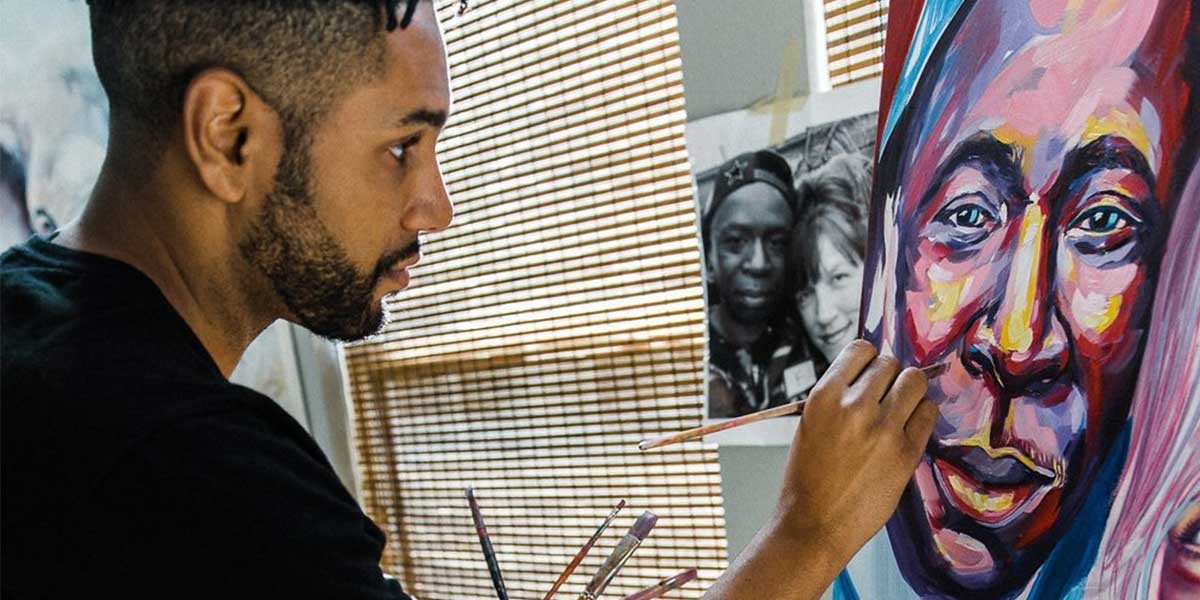 A man painting a colorful portrait of a face on a vertical canvas indoors near a window with blinds.