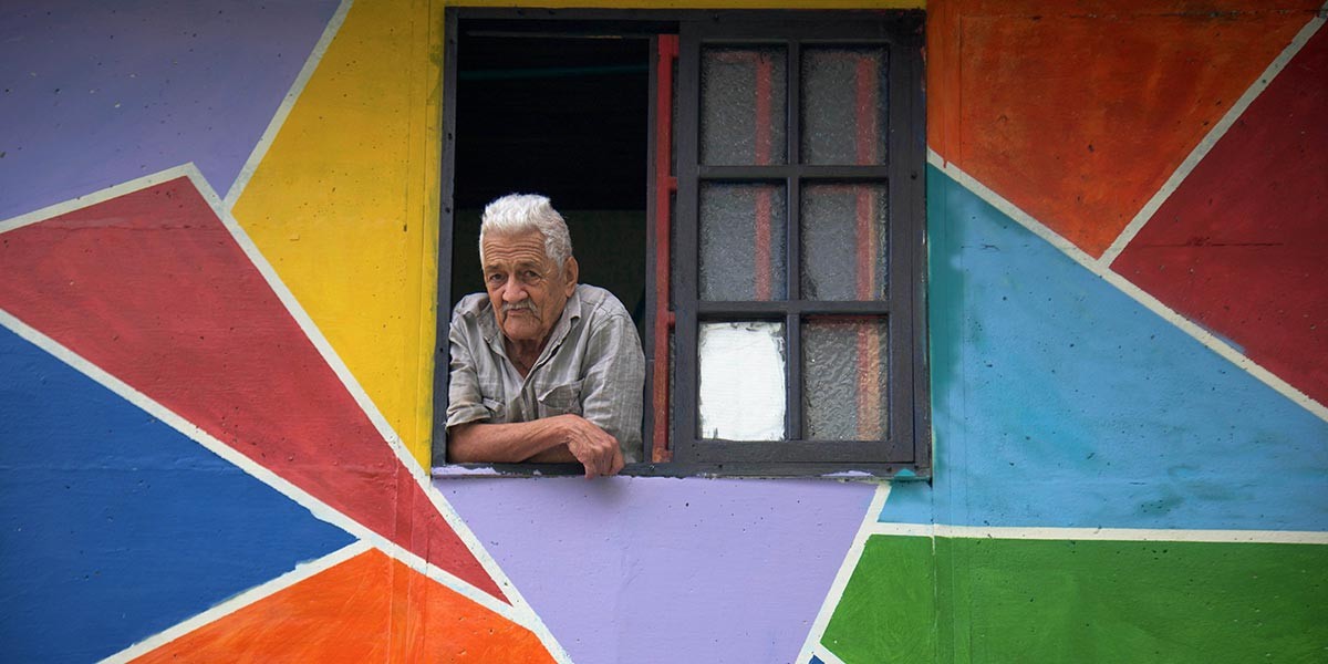 Elderly man leaning on a window sill of a colorful geometric-patterned wall.