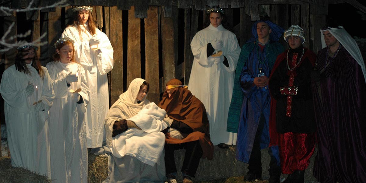 Group dressed as nativity characters, including Mary, Joseph, angels, and wise men in a wooden stable.
