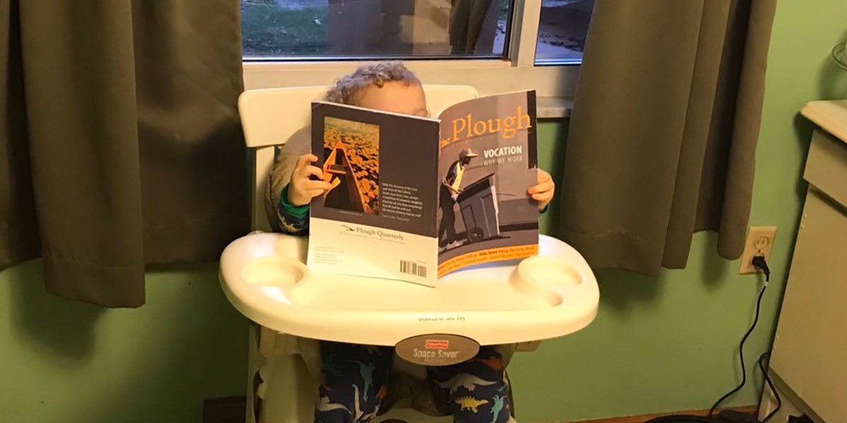 Toddler sitting in high chair reading a Plough Quarterly magazine with dinosaur pajamas.