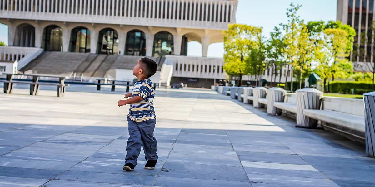 Young boy in striped shirt walking on paved plaza near benches and green trees in a city setting.