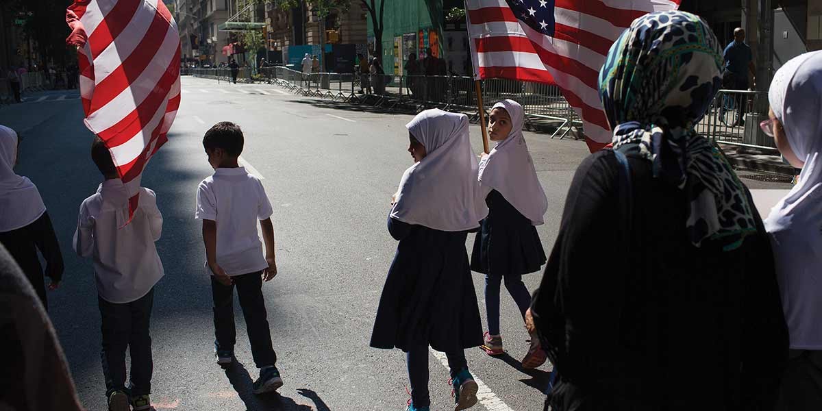 Children and a woman wearing hijabs walking on a street holding American flags in daylight.