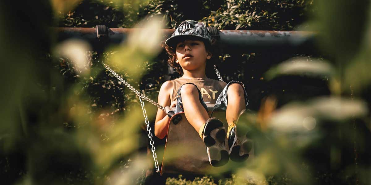 Child wearing a cap swinging outdoors, partially seen through blurred green foliage.