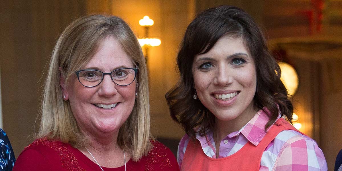 Two smiling women standing side by side indoors, one with glasses and blonde hair, the other with dark hair.