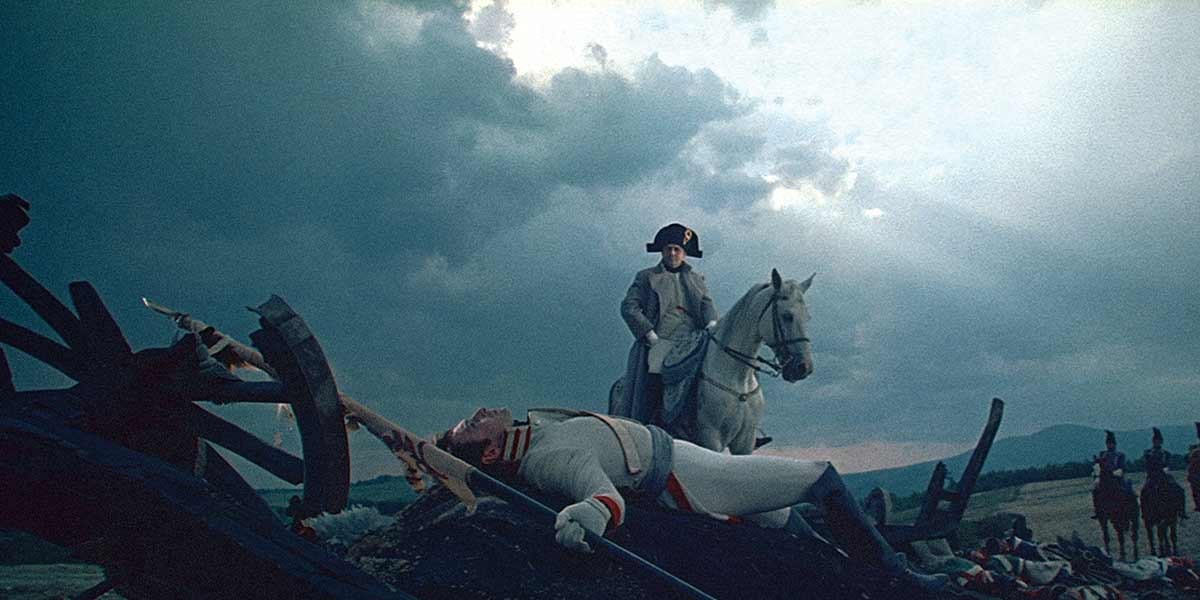 Napoleon Bonaparte in uniform on a white horse overlooking a fallen soldier amid a battlefield scene.