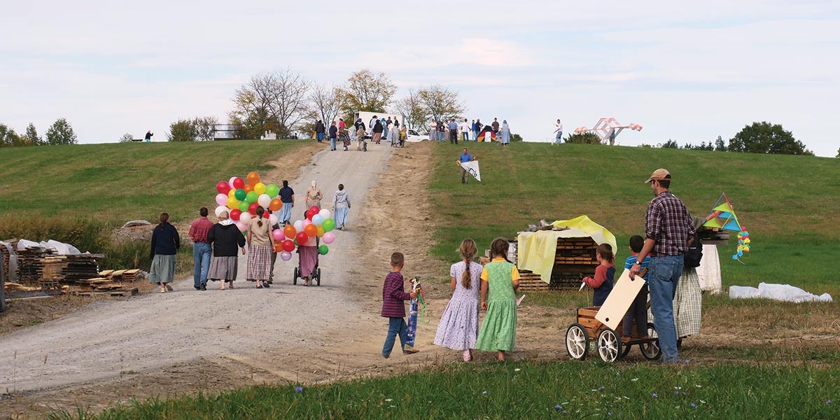 Group of children and adults walking up a grassy hill, some holding colorful balloons and kites, outdoors.