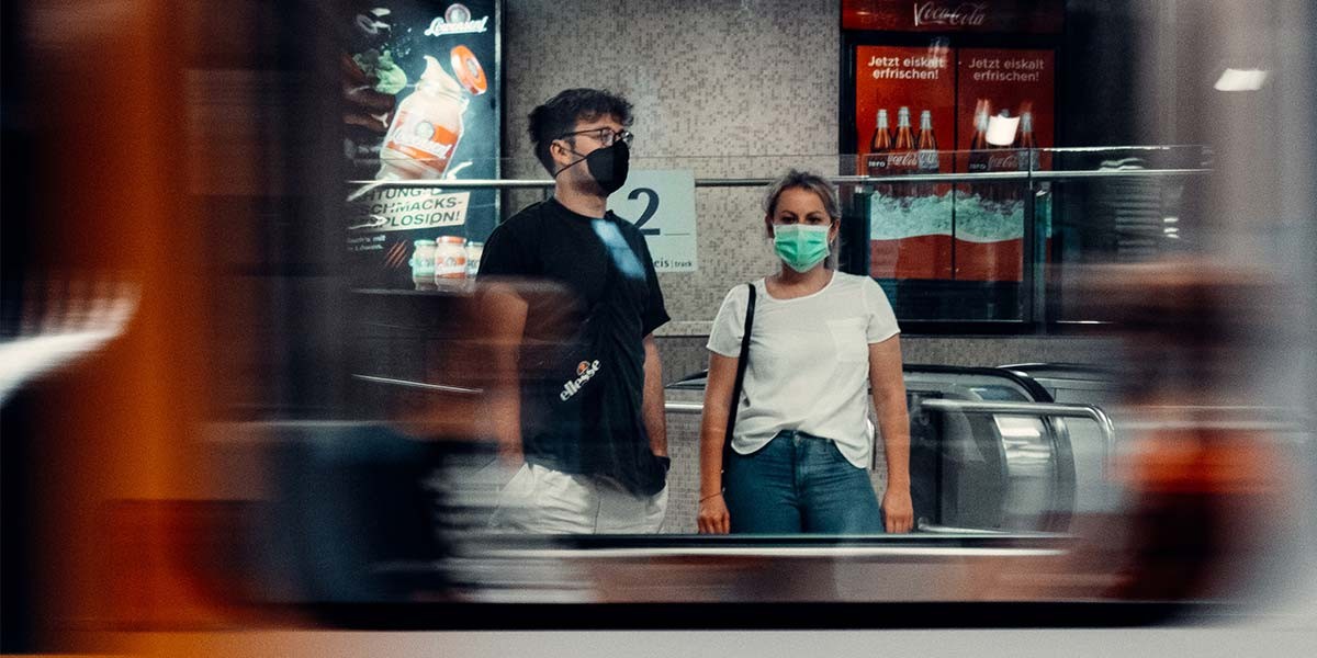 Man in black shirt and woman in white shirt wearing masks standing near escalator with blurred train passing by.