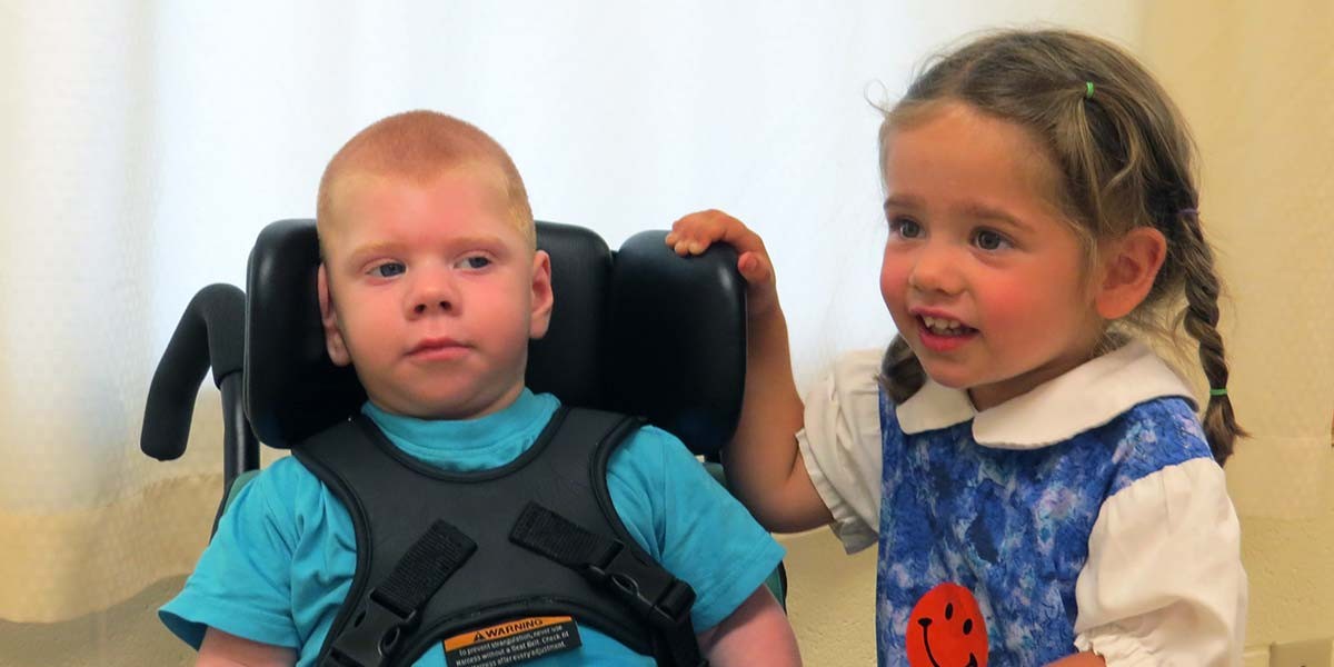 Young girl with braids smiling and standing next to a boy in a blue shirt seated in a wheelchair.