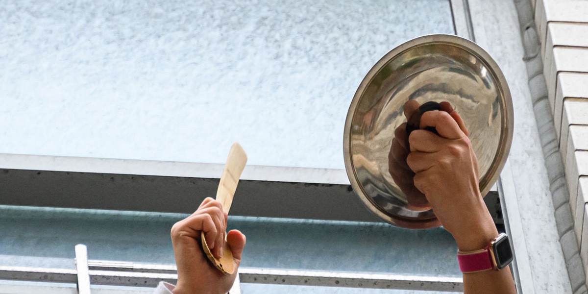 Two hands holding a spoon and a metal pot lid raised against a window.