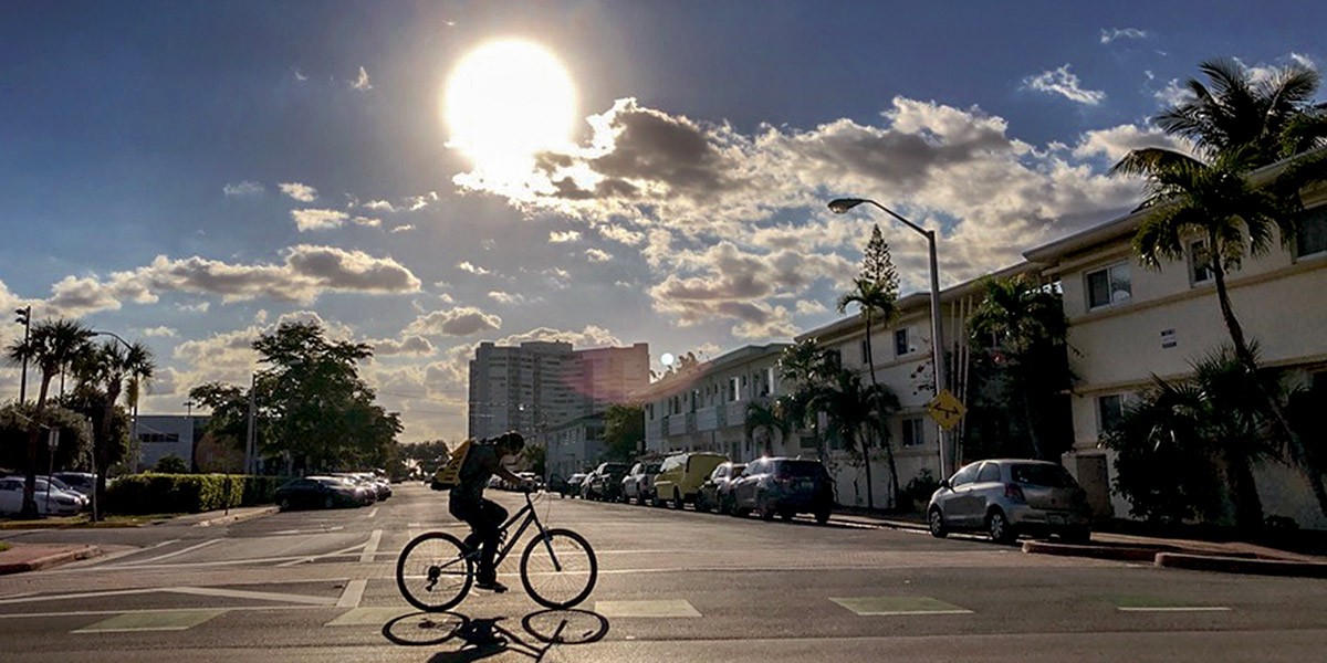 Person riding a bicycle across a city street with sun shining through partly cloudy sky.