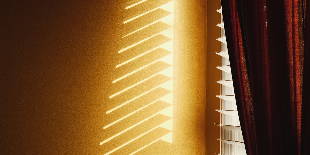 Sunlight casting geometric shadows from window blinds onto a yellow wall beside a dark curtain.