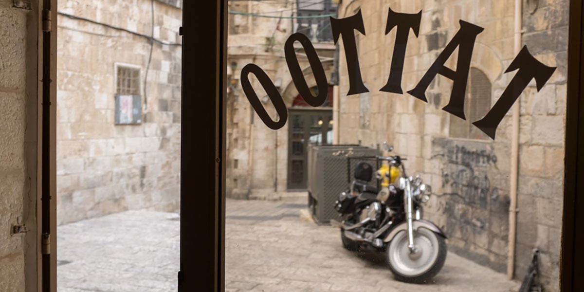 View through a window with the word 'TATTOO' reversed, showing a parked motorcycle outside on stone pavement.
