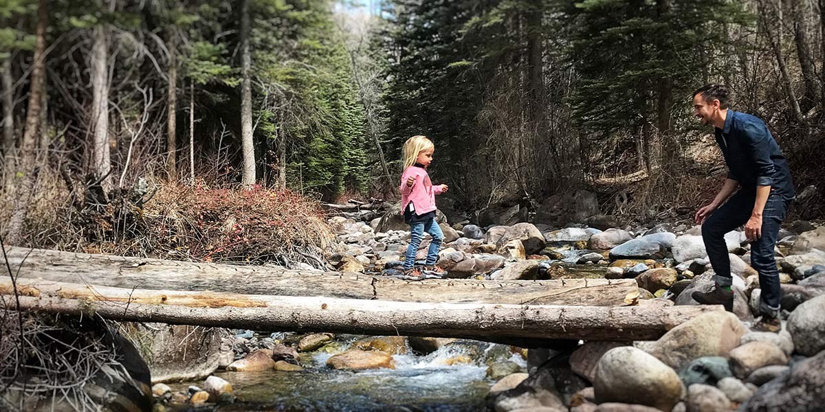Child in pink jacket crossing stream on log with adult man watching in forested rocky area.