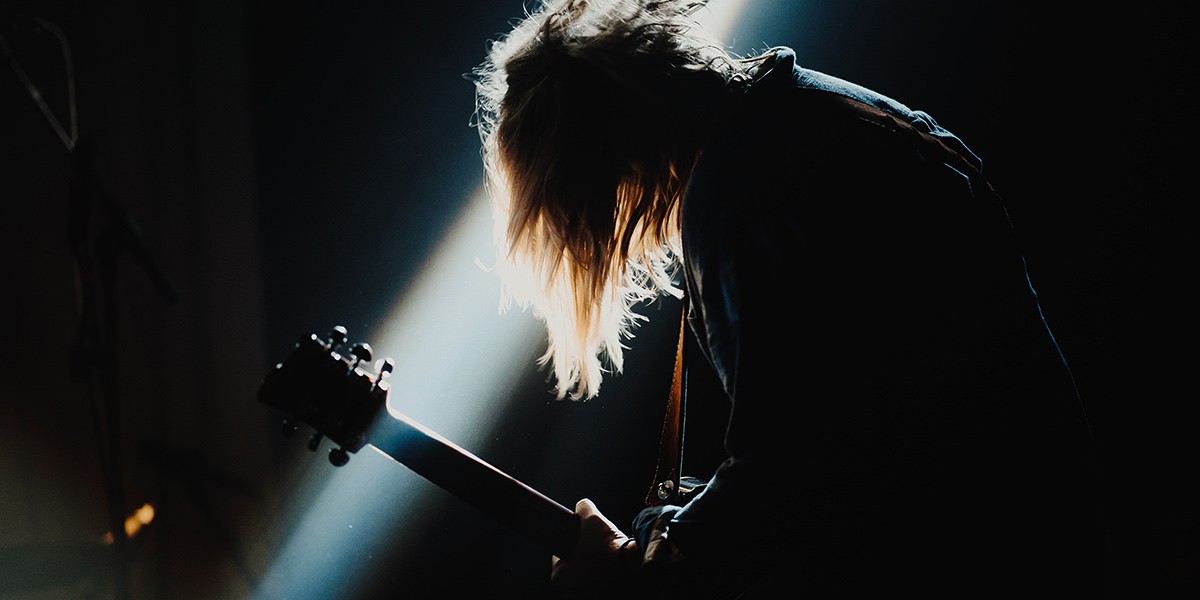 Silhouetted musician playing electric guitar with light shining on their hair and instrument.