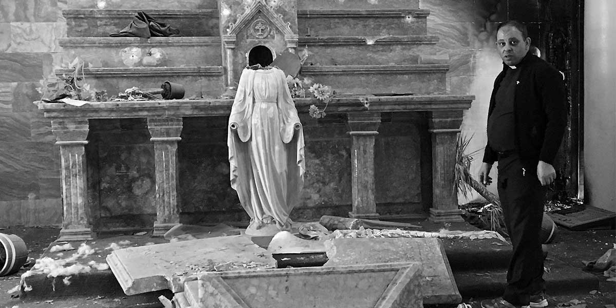 Man in dark clothes stands beside damaged altar with headless statue in a ruined church.