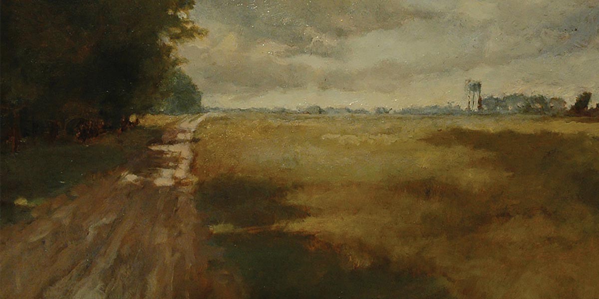 A rural dirt road beside a field under a cloudy, overcast sky with trees and a distant water tower.