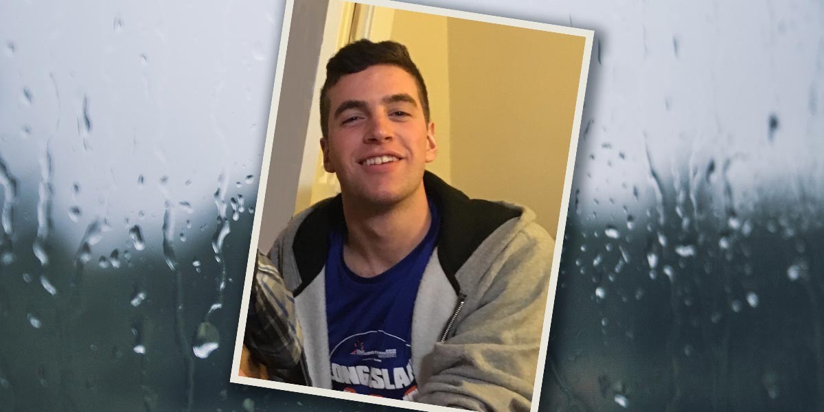 Young man smiling wearing a blue shirt and grey hoodie indoors with a beige wall background.