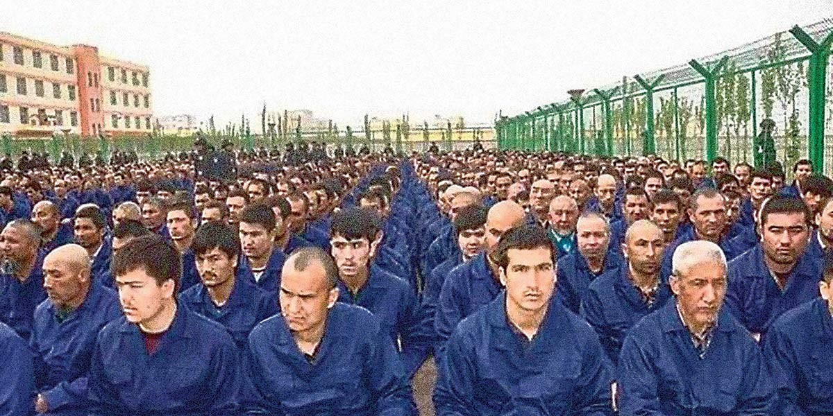 Large group of men wearing blue uniforms sitting outdoors in rows beside a fence and buildings.
