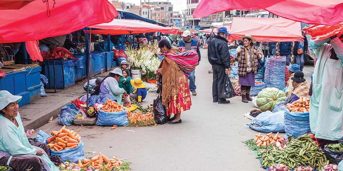 People shopping and selling fresh produce under red tents at a bustling outdoor market.