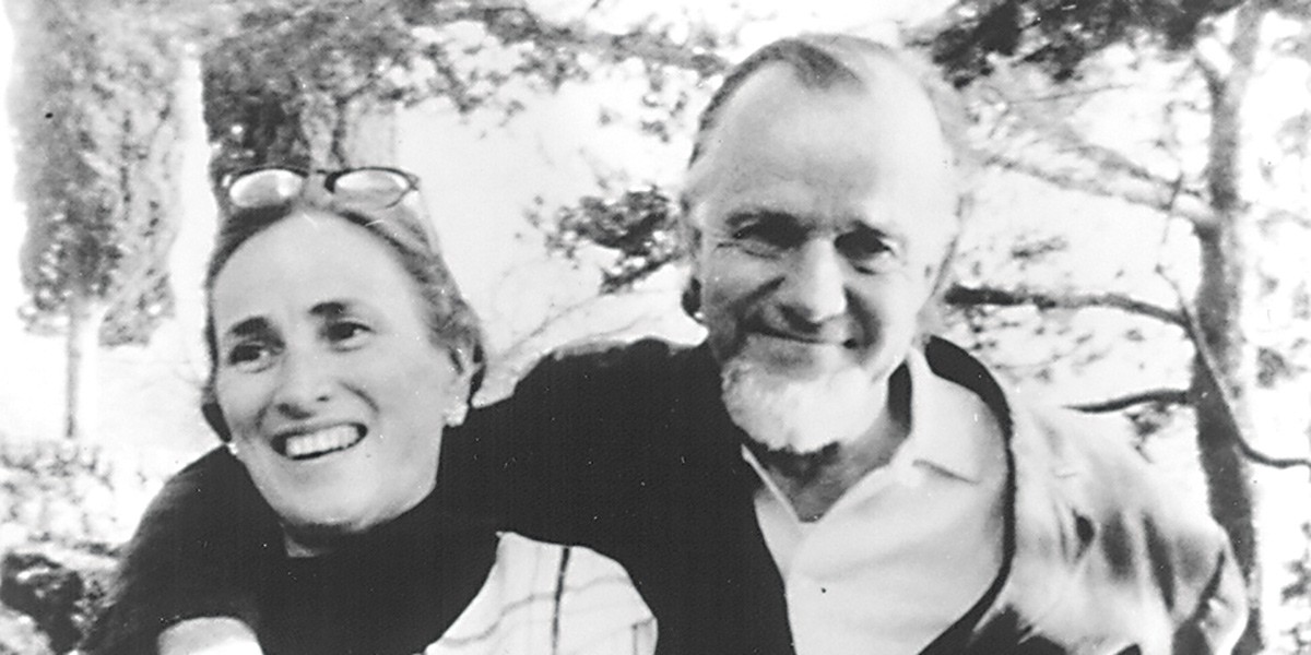 Black and white photo of an older man and woman smiling outdoors with trees in the background.