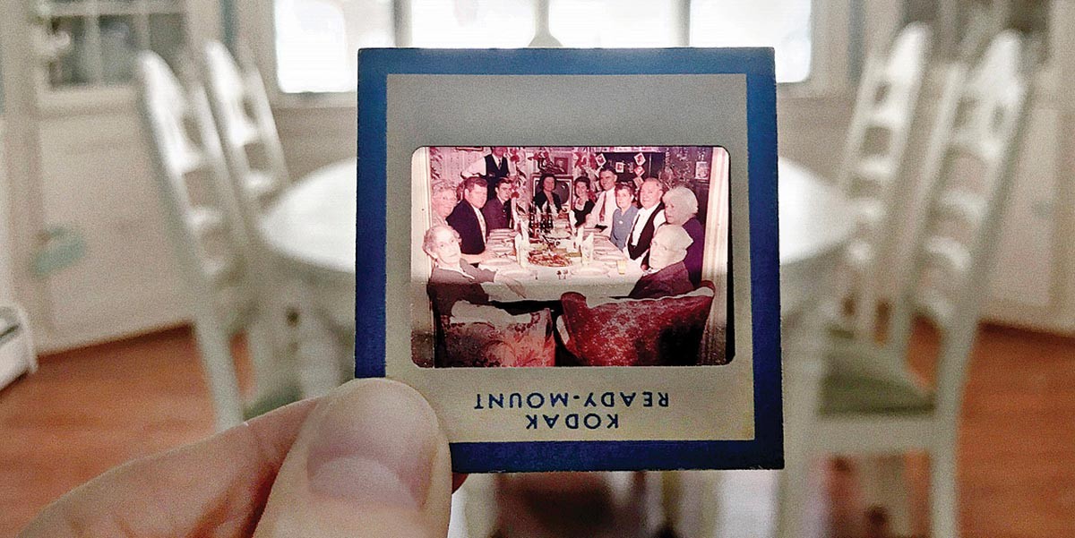 Hand holding a vintage Kodak slide showing a group of people seated around a dining table.