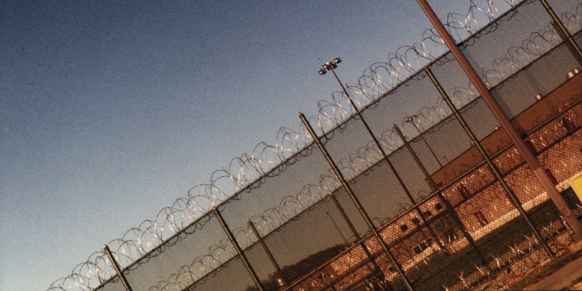 Barbed wire fence topped with coils surrounding a prison building under blue sky.