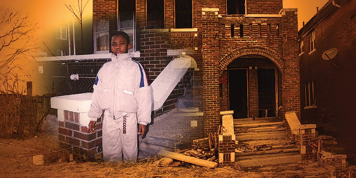 Young boy in light jacket and pants standing in front of a damaged, abandoned brick house at dusk.